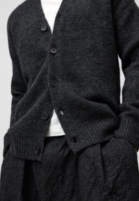 Koldo i fårets ull med V-ringning, fem svarta knappar, ribbade ärmslut och en något lös passform, parat med strukturerade svarta byxor.