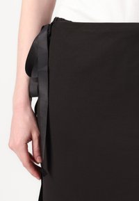 Melna kokvilnas priekšauts ar gludu tekstūru, ar sānu lentu, lai pielāgotu izmēru, un vienkāršu, minimālistisku dizainu.