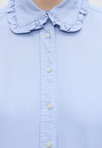 Chemise bleu clair avec col à volants et cinq boutons blancs au centre devant, montrant la partie supérieure de la poitrine sans manches.