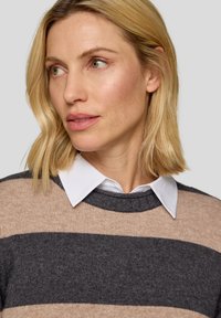 Beige und anthrazit gestreifter Pullover aus weichem Stoff mit rundem Ausschnitt. Darunter wird ein weißes Hemd mit Kragen getragen.