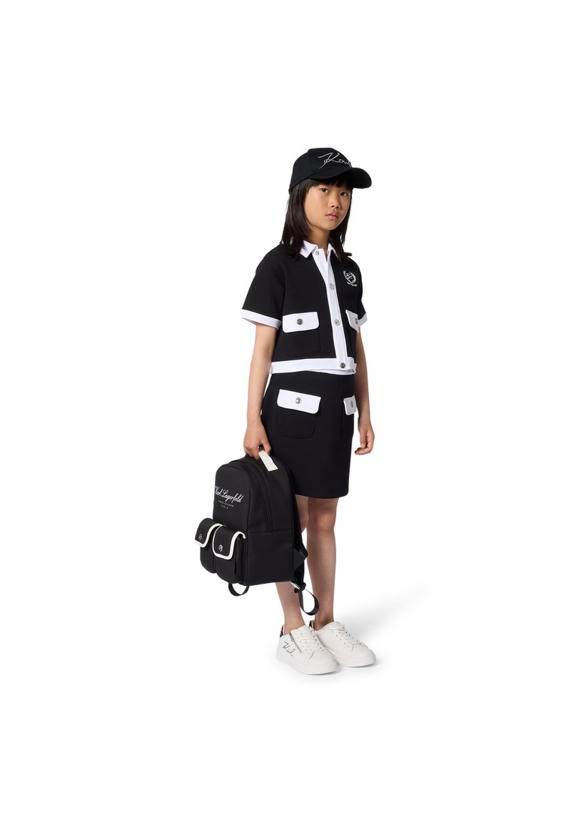 Σετ κοντομάνικου πουκάμισου και φούστας σε ασπρόμαυρο με τσέπες· κορίτσι φοράει μαύρο καπέλο, κρατάει μαύρο σακίδιο με λευκές λεπτομέρειες και παπούτσια.
