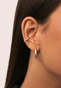 Oreja de mujer con cabello oscuro y lacio, lleva un aro de plata retorcido en la parte superior de la oreja y un pendiente de plata lisa en el lóbulo de la oreja.