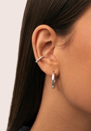 Oreja de mujer con cabello oscuro y lacio, lleva un aro de plata retorcido en la parte superior de la oreja y un pendiente de plata lisa en el lóbulo de la oreja.