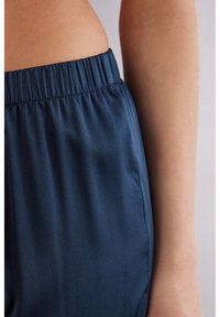 Elastische taille donkerblauwe broek met een gladde textuur, met een rechte pijp en zijzakken, voor een deel zichtbaar tegen een huidskleurige achtergrond.