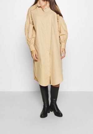 Mujer vestida con un vestido midi beige de manga larga con botones y mangas abullonadas, y botas negras hasta la rodilla, de pie sobre un suelo gris.