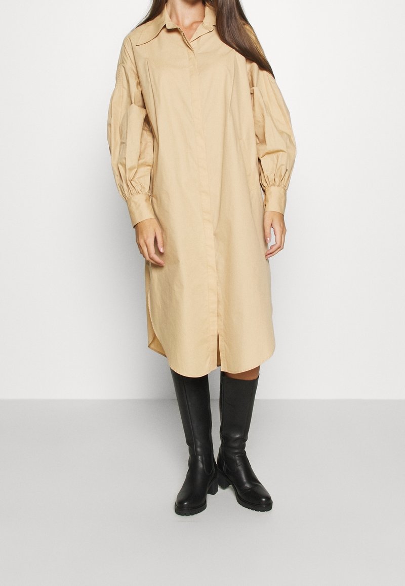 Femme portant une robe midi beige à manches longues boutonnée avec des manches bouffantes et des bottes noires montantes jusqu'aux genoux, debout sur un sol gris.
