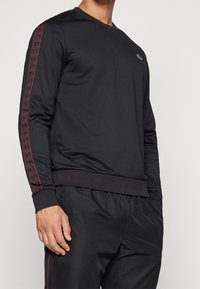 Svart långärmad träningsskjorta i fuktavvisande material, med röda geometriska mönster på ärmarna, ribbade ärmslut och logotyp på bröstet.