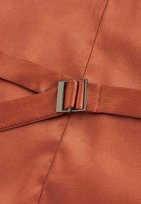 Tissu satin couleur rouille avec une texture lisse ; comporte une boucle en métal et un détail de sangle repliée sur un côté.