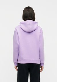 Άτομο με ίσια καστανά μαλλιά στέκεται με την πλάτη γυρισμένη φορώντας μια παστέλ μοβ φούτερ με κουκούλα και μαύρο παντελόνι μπροστά σε απλό φόντο.