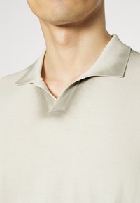 Polo tričko v světle béžové bavlně, s klasickým límcem, měkkou strukturou a uvolněným střihem. Vyznačuje se minimalistickým designem.