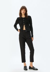Zwarte geribde cardigan met ronde hals en knoopsluiting, gecombineerd met hoge zwarte broeken en glanzende zwarte loafers.