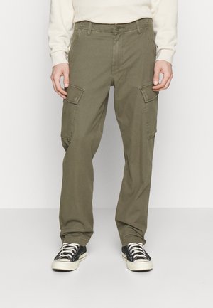 Mand iført olivengrøn cargo-bukser med klaffelommer og sorte lave sneakers med hvide snørebånd og såler, stående på et hvidt gulv.