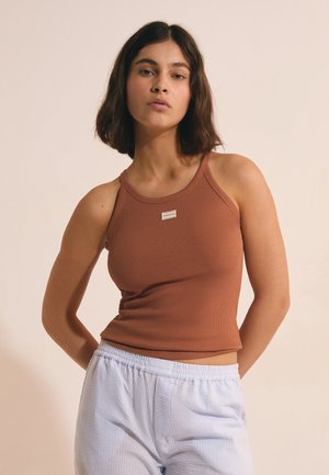 Joven mujer con cabello corto oscuro que lleva una camiseta sin mangas marrón acanalada y pantalones texturizados azul claro sobre un fondo beige.