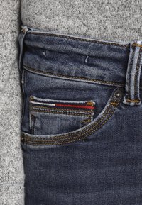 Vaqueros de denim azul oscuro con un pequeño bolsillo frontal que presenta rayas rojas y amarillas. Se aprecian costuras en contraste y una textura envejecida.