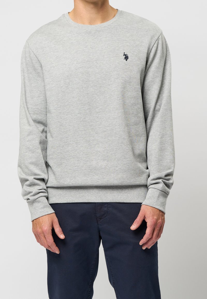 Homme portant un sweat-shirt gris clair à manches longues avec un petit logo noir sur la poitrine et un pantalon bleu foncé, debout sur fond blanc.