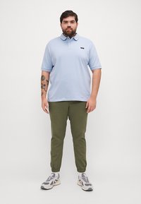 Jasnoniebieska koszulka polo z kołnierzykiem, zestawiona z oliwkowymi spodniami dresowymi. Stylizacja zawiera akcent w postaci logo oraz szare sneakersy.
