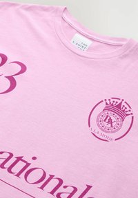 Rozā kokvilnas T-krekls ar tumši rozā grafiku, tostarp logo ar kroni un tekstu. Apaļš kakls, īsas piedurknes, maiga tekstūra.