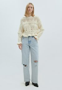 Cremfarbener Strickpullover mit offenen Muster, langen Ärmeln und rüschendem Saum. Zartblaue Röhrenjeans. Schwarze spitz zulaufende Schuhe.