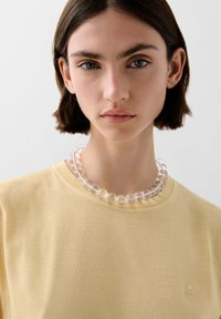 Eine klare Perlenkette liegt auf einem hellgelben Rundhals-T-Shirt, das glatte, semi-transparente Perlen mit subtilen Farbvariationen zeigt.