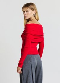 Roter Schulterfreier Pullover mit gerippter Struktur und langen Ärmeln, kombiniert mit grauen Faltenhosen. Der Pullover hat einen drapierten Ausschnitt.