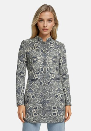 Blond vrouw die een langermige, geknoopte jas draagt met een gedetailleerd marineblauw-wit bloemenpatroon en staat tegen een effen achtergrond.