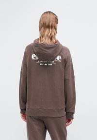 Osoba z falującymi, blond włosami do ramion, ubrana w brązową bluzę z kapturem i dopasowane spodnie, stoi tyłem, pokazując napis "ellesse Perugia Italia" na plecach.