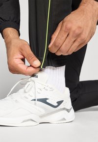 Białe sportowe buty z szarym logo, w połączeniu z czarnymi spodniami dresowymi z neonowo zielonym wykończeniem. Ręce poprawiają brzeg spodni.