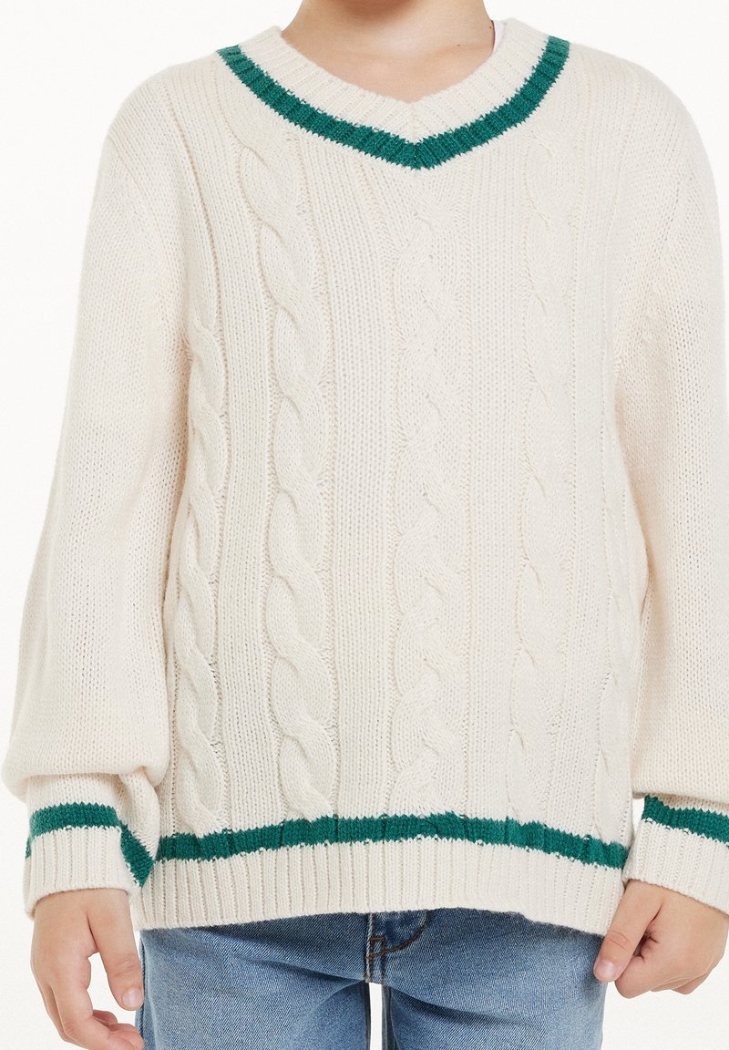 Enfant portant un pull torsadé crème avec des rayures vertes au col en V, aux poignets et à l’ourlet, porté avec un jean bleu.