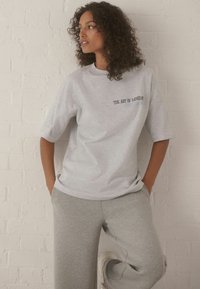 Graues Baumwoll-T-Shirt mit kurzen Ärmeln, das den Text "DIE KUNST DER BEWEGUNG" in dunkler Schrift zeigt. Kombiniert mit grauen Jogginghosen vor einer weißen Wand.