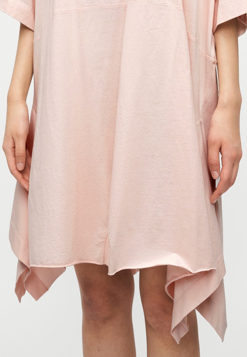 Robe rose ample en coton doux, avec un ourlet haut-bas et des poches latérales. Texture lisse avec un design minimaliste.