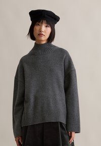 Γυναίκα με κοντά μαύρα μαλλιά, φορώντας μαύρο μπερέ, σκούρο γκρι ριμπ τσιρκωτό πουλόβερ και μαύρη πλισέ φούστα, που στέκεται απέναντι από έναν απλό τοίχο.