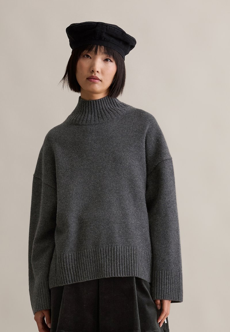 Γυναίκα με κοντά μαύρα μαλλιά, φορώντας μαύρο μπερέ, σκούρο γκρι ριμπ τσιρκωτό πουλόβερ και μαύρη πλισέ φούστα, που στέκεται απέναντι από έναν απλό τοίχο.