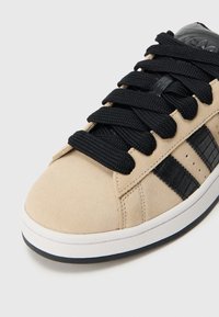 Μπεζ σουέντ αθλητικά παπούτσια με μαύρες συνθετικές δερμάτινες λεπτομέρειες, χοντρές μαύρες κορδόνια και λευκή ελαστική σόλα. Διαθέτουν τρεις μαύρες ρίγες στο πλάι.