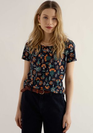 Tânără cu păr blond ondulat, purtând un top negru cu mâneci scurte și modele florale, pantaloni închiși la culoare și o curea maro împletită.