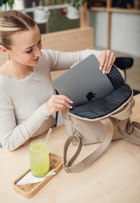 Beige leather backpack with a zippered closure, featuring a patterned black interior. A woman places a laptop inside. Green drink nearby.