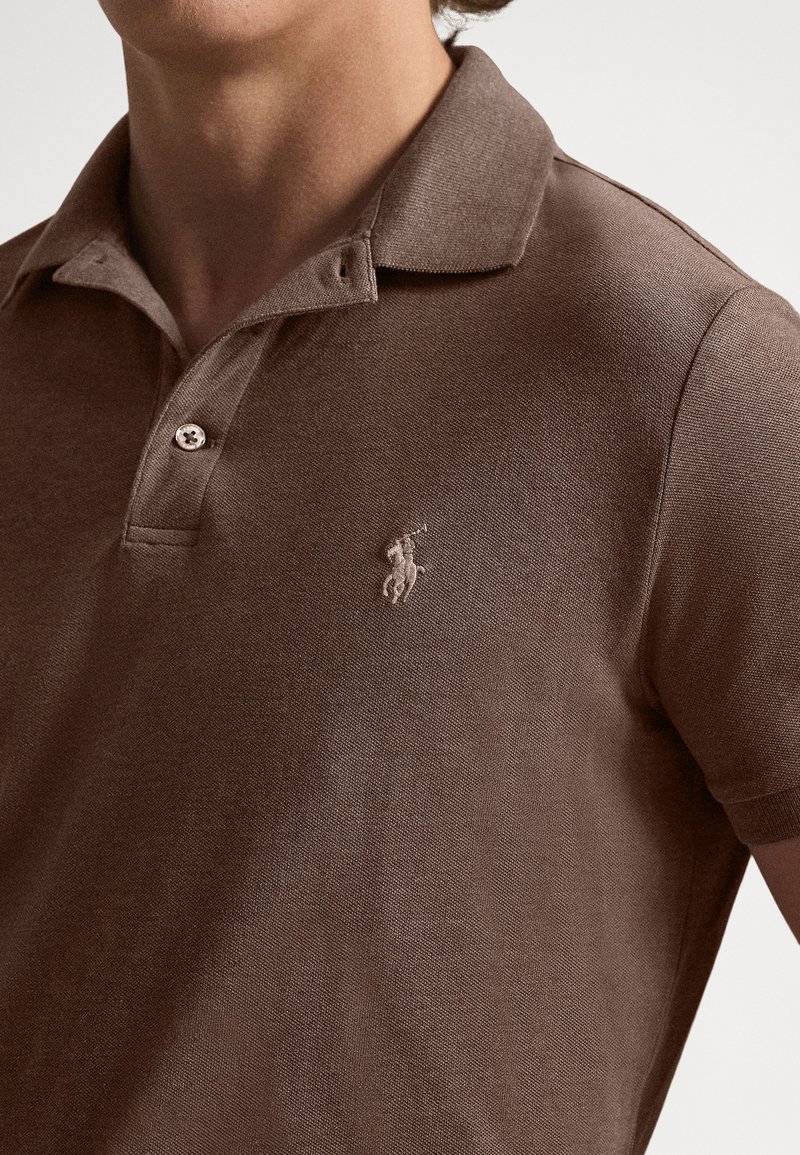 Nahaufnahme einer Person, die ein braunes Poloshirt mit Kragen, zwei Knöpfen und einem kleinen bestickten Logo eines Reiters auf einem Pferd auf der Brust trägt.