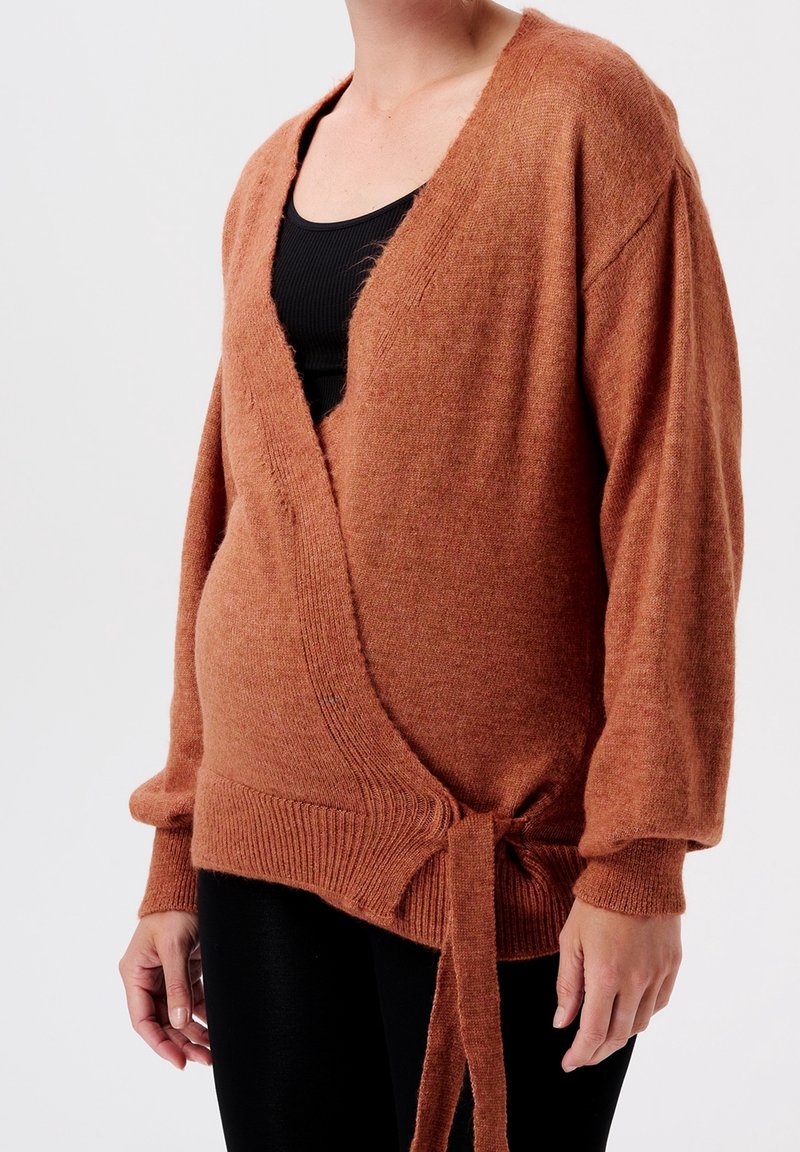 Personne portant un pull cache-cœur couleur rouille noué à la taille sur un haut noir sans manches, debout devant un fond uni.