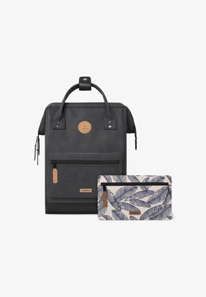 Mochila negra texturizada con un asa cosida, un bolsillo frontal y una bolsa de accesorio de color crema con un patrón de hojas. Presenta una etiqueta con el logotipo.