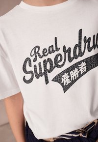 Vitt t-shirt med svart texttryck i sprucken stil som lyder "Real Superdry" och japanska tecken, bärs med ett blått randigt bälte och beige byxor.