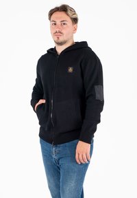 Felpa nera con zip, tasca frontale, polsini a costine e una piccola etichetta con logo. Il tessuto appare morbido e con una texture, con design con cappuccio.