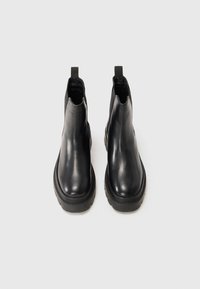 Een paar zwarte leren Chelsea-boots met stevige zolen en trekstrips, gezien van voren op een lichtgrijze achtergrond.