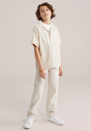 Béžová krátky rukáv košeľa s pohodlným strihom, doplnená voľnými béžovými nohavicami. Oblečenie dotvárajú biele tenisky a drevený náramok.