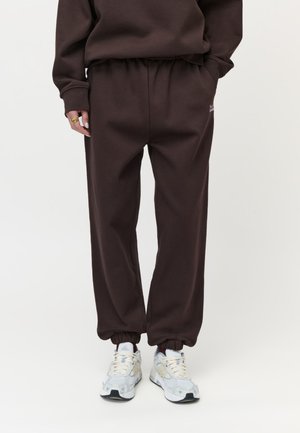 Person iført mørkebrune sweatpants med elastiske manchetter og hvide sportssko, stående mod en ensfarvet lys baggrund.