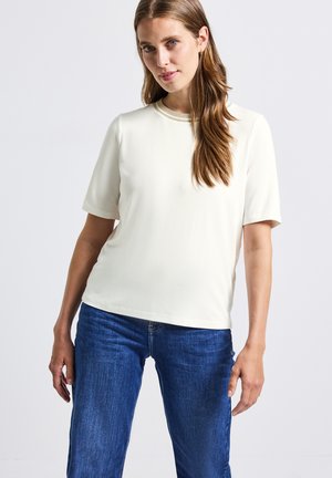 Femme aux longs cheveux bruns portant une chemise blanche à manches courtes et un jean bleu, se tenant devant un fond clair uni.