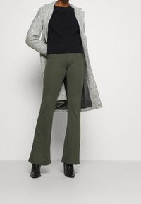 Persona che indossa pantaloni a zampa d'elefante verde oliva, stivali neri alla caviglia, maglione nero a maglia e un lungo cappotto grigio con le mani nelle tasche.