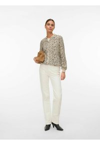 Femme portant une veste zippée beige et noire à motifs, pantalon blanc, bottines noires, tenant une pochette marron, debout sur fond blanc.