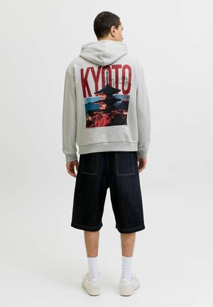 Bărbat cu fața spre spate, purtând hanorac gri cu imprimeu "KYOTO" și pagodă, pantaloni scurți negri până la genunchi, șosete albe și adidași pe un fundal simplu.