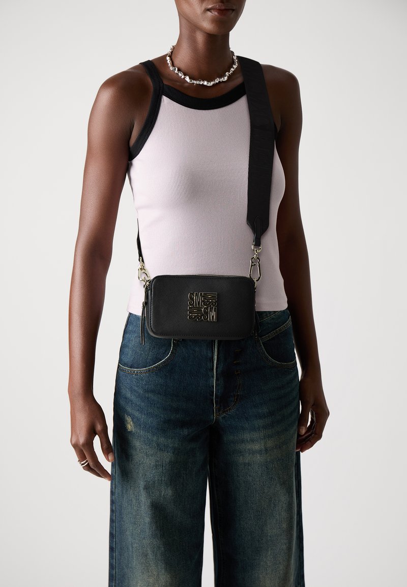 Sort læder crossbody taske med logo detalje, fastgjort skulderrem og lynlås. Båret over en lys tanktop og løstsiddende jeans.