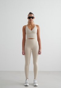 Beige sportbeha en high-waisted leggings van glad materiaal. Gekocht met witte en grijze sportschoenen en donkere zonnebril.