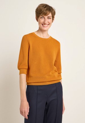 Mustard yellow knit sweater with three-quarter sleeves, round neckline, and textured fabric. Paired with dark navy trousers.